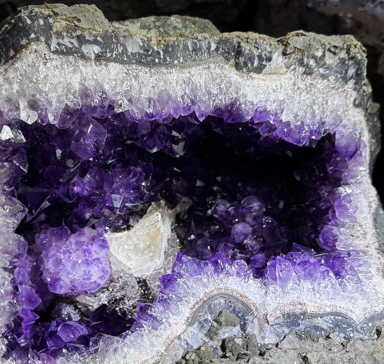 An open amethyst geode seen in cross-section: outer basalt, a band of bluish-gray agate, quartz crystal, violet amethyst toward the hollow and a white calcite crystal