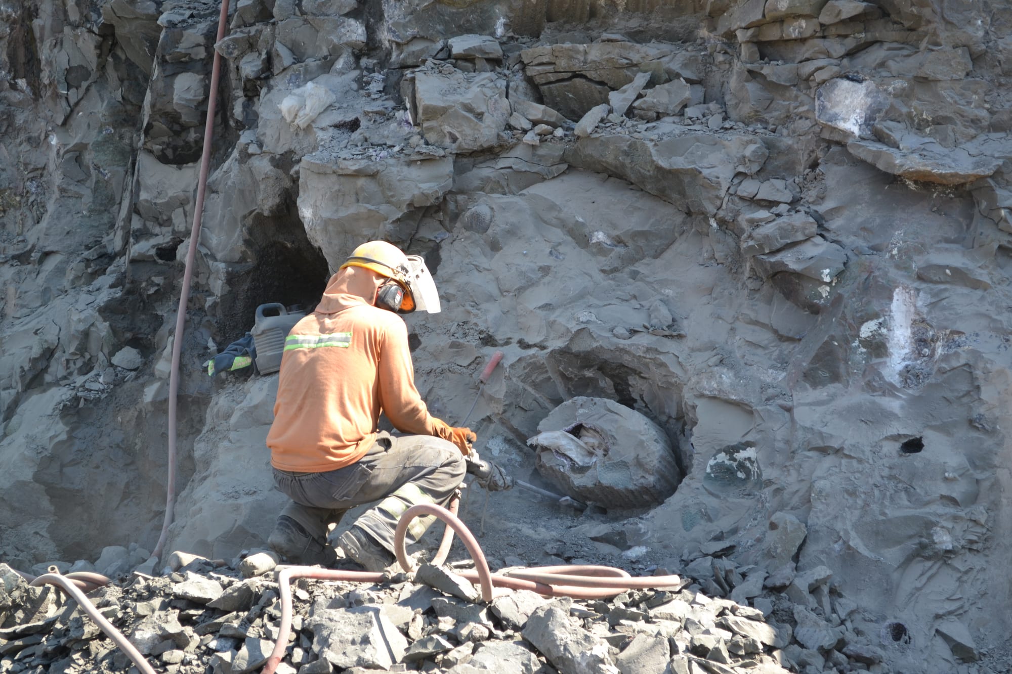 A hand miner in a helmet and face shield works with a pneumatic hammer beside a geode at the quarry face